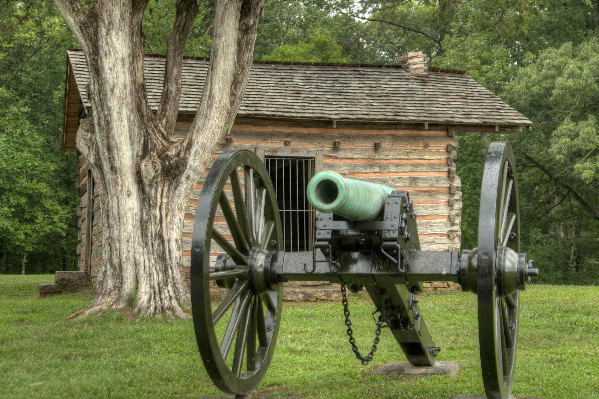 Cannon sitting in a green field in front a rustic log cabin